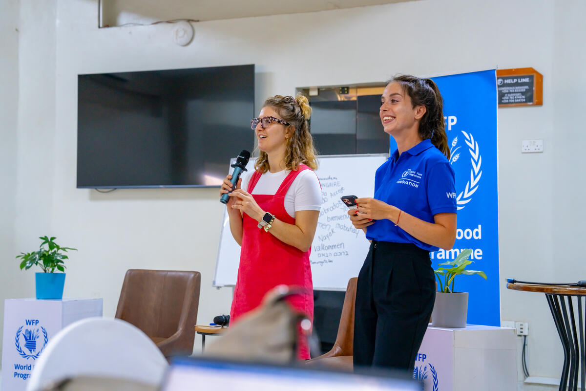 Two women stand with microphones, speaking off camera.