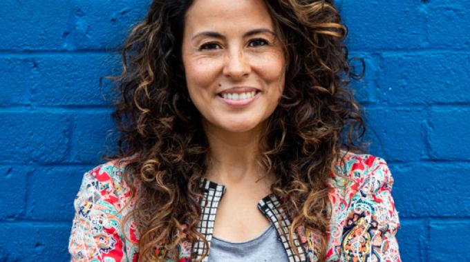 Photo of Andrea Camargo against a blue brick background smiling.