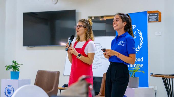 Two women stand with microphones, speaking off camera.
